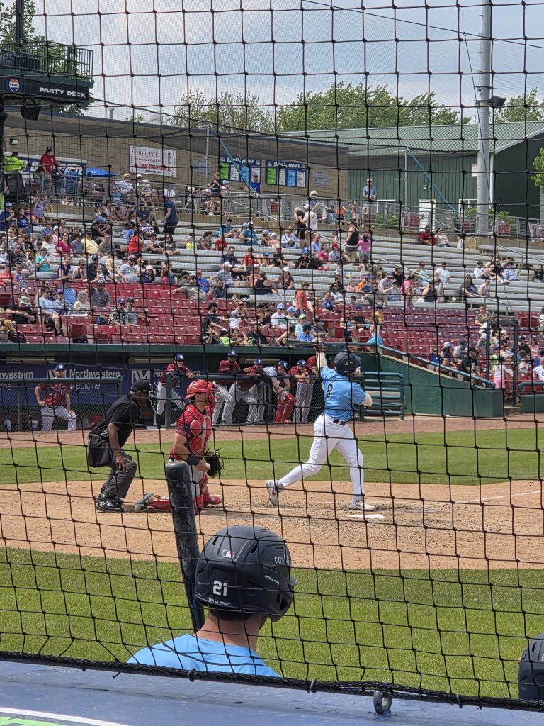 View from behind home plate at Northwestern Medicine Field, Kane County Cougars game May 2026, batter at the plate with catcher and umpire, crowd visible in the stands on a sunny afternoon
